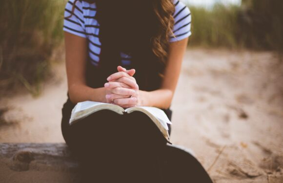 A woman sitting outdoors on a wooden log, with hands clasped in prayer over an open Bible, on a sandy area with tall grass in the background.