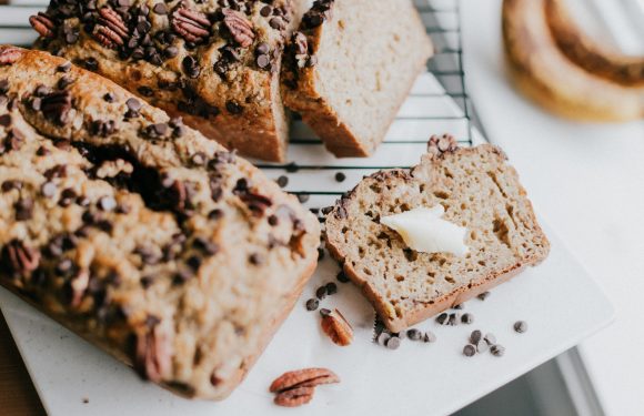 Banana bread with chocolate chips and pecans, with a slice topped with butter on a white cutting board. A bagel is partially visible in the background.