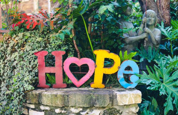 Colorful metal letters spelling "HOPE" with a pink heart in place of the letter "O" are displayed on a stone ledge surrounded by lush green plants and garden statues.