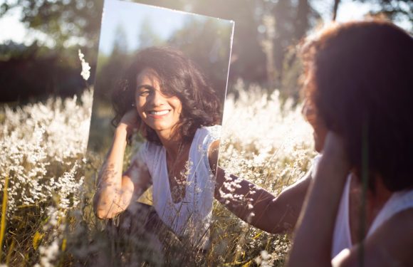 A woman holding a mirror outdoors in a field of white flowers, smiling as she looks at her reflection, with sunlight and trees in the background.