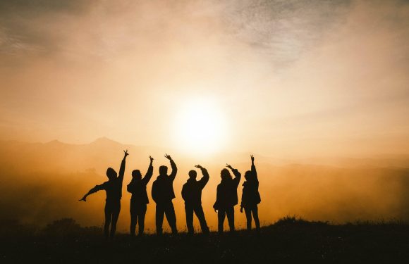 Six people in uniform pose with peace signs during sunset, standing on a hill with a blurred landscape and mountains in the background.