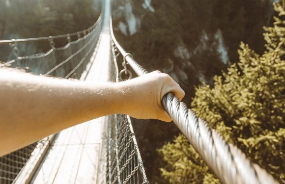 Looking down a suspension bridge over a forested canyon, with a person's hand holding onto the metal cable rail and sunlight shining from the top left.