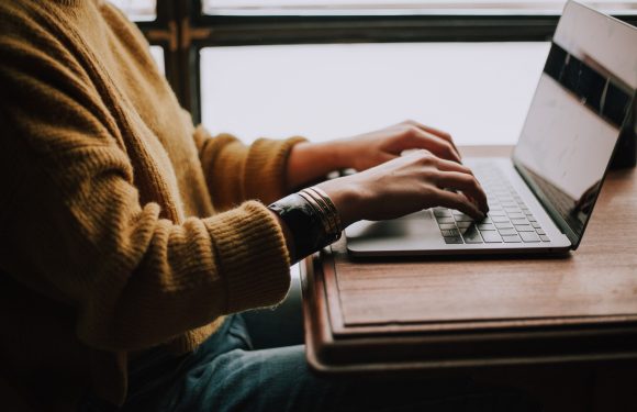 Person wearing a striped bracelet using a laptop at a wooden table near a window, with light coming in from outside.