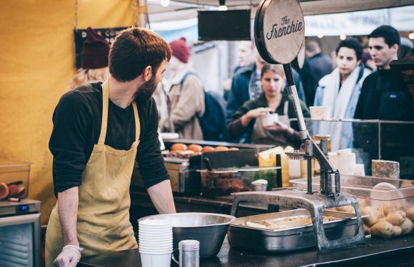 A man in a black shirt and beige apron works at a street food stall with a crowd of people behind him, offering baked potatoes and other foods.