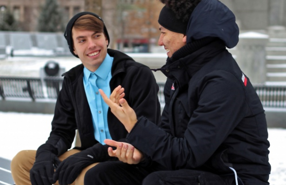 Two young men sit on a bench outdoors in winter, smiling and talking, dressed warmly with one in a uniform and the other in a hoodie and winter gear.