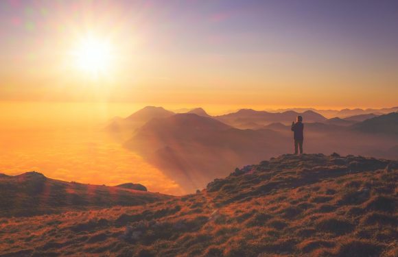 A person standing on a grassy mountain ridge, overlooking a mountain range with clouds below and a bright sun in the sky during sunset or sunrise.