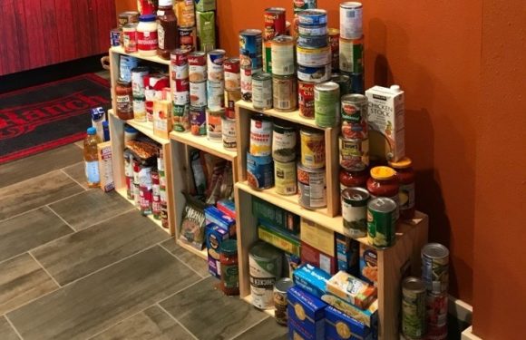 Wooden shelves filled with canned and boxed food items, placed against an orange wall in a room with tiled flooring.