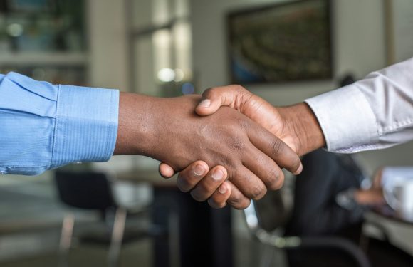 A close-up of two people shaking hands, one wearing a light blue shirt and the other in a white shirt, in a professional setting with a blurred background.
