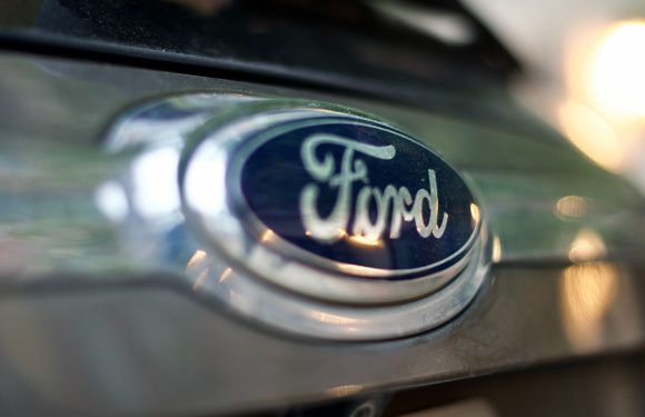Close-up of a Ford emblem on a vehicle's front grille with a blurred background and sunlight reflecting off the chrome surface.