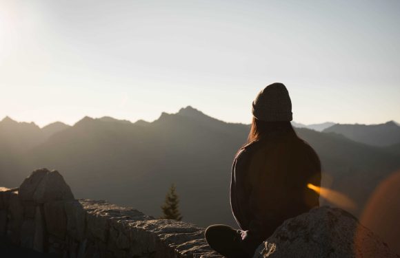 A woman sitting on a stone wall, overlooking mountains at sunset, wearing a beanie and jacket, with her back to the camera.