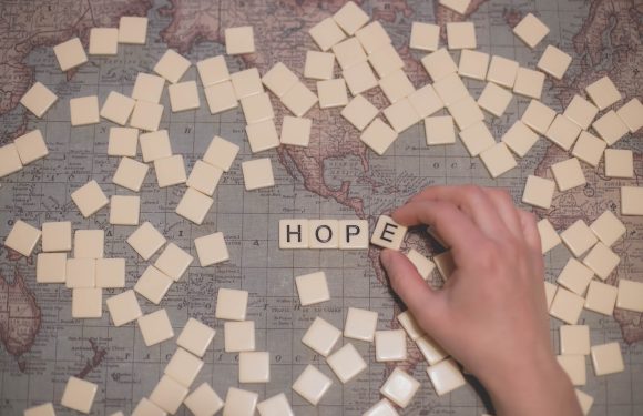 A hand arranging tiles on a world map with the tiles spelling "HOPE" amidst scattered white tiles.