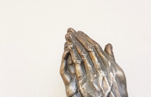 A metallic sculpture of hands in prayer position, with intricate details on the fingers and knuckles, set against a plain wall background.