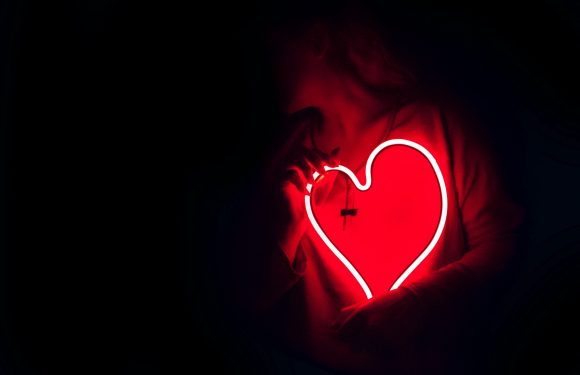 A person in a red hoodie holds a glowing heart-shaped light with a crack running through it, set against a dark background.