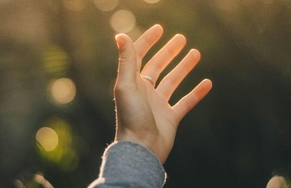 A person's hand with a ring on the ring finger reaching upward towards the bright, bokeh-filled sky during sunset, wearing a gray sleeve.