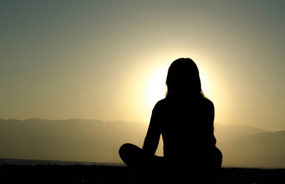 A person sitting cross-legged on the ground, facing a sunset over distant mountains, with their silhouette visible against the bright sky.