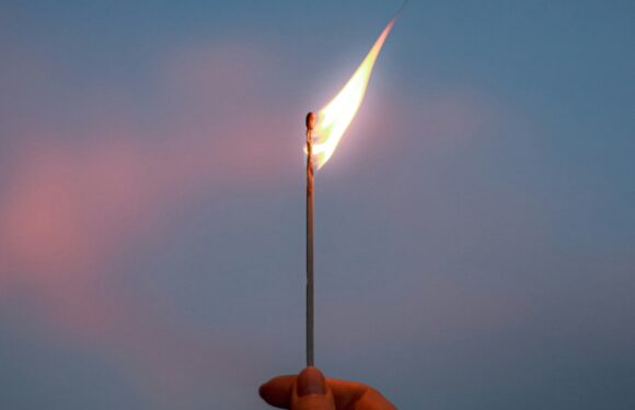 A hand holds a lit match with a bright flame against a twilight sky. The background is mostly blue with soft pinkish clouds.