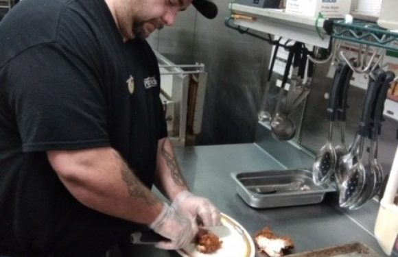 Man in a black uniform and cap prepares food on a plate in a commercial kitchen with baking trays and utensils around him.