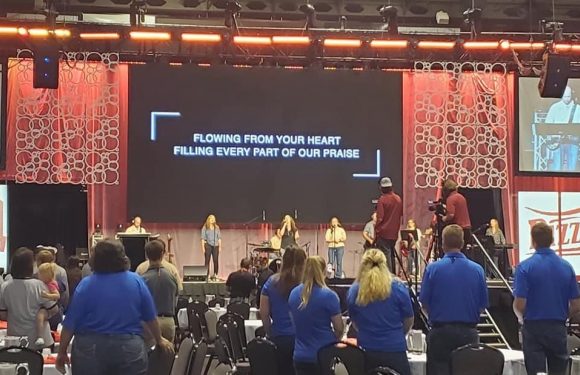 People gather in a large indoor venue with a stage, where a worship band performs. Some attendees wear blue shirts, possibly staff or volunteers.
