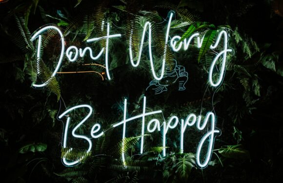 A neon sign with the message "Don't Worry Be Happy" surrounded by lush green tropical plants.