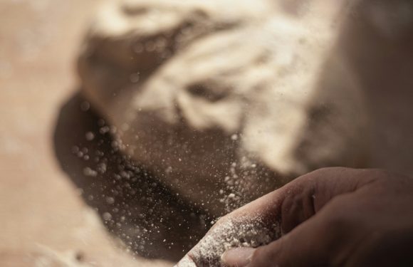 A hand dusted with flour touches a surface with a dusting of flour and a partially shaped dough or clay sculpture in the background.