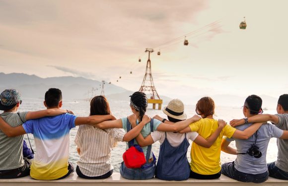 Eight friends sit on a bench with arms around each other, facing water and a sunset, with mountains and cable cars in the background.