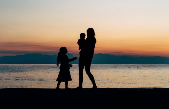 Silhouettes of a woman and two children by the water at sunset, with mountains in the background. The woman is holding a young child, and the other child interacts with her.