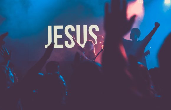 People raise their hands in worship or praise in front of a large screen displaying the word "JESUS" during a church service or religious event.