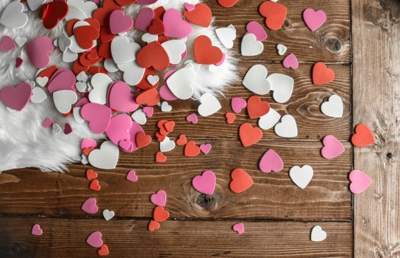 Colorful heart-shaped confetti in pink, red, and white spread across a wooden surface with a fluffy white fabric in the top left corner.