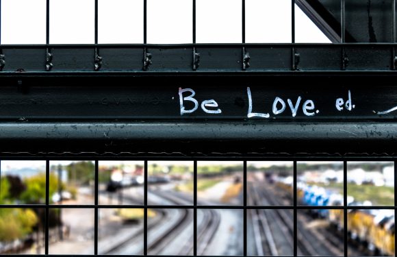 A close-up of a metal fence with graffiti reading "Be Love." Overhead train tracks and a blurred parking lot are visible in the background.