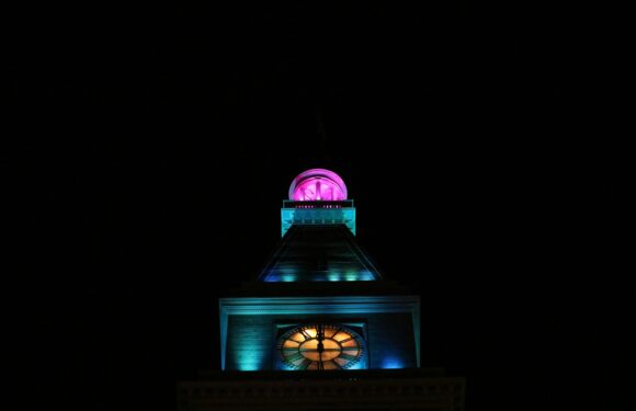 A lit clock tower at night features colorful lighting, with a pink dome at the top, a blue section below, and a glowing clock face near the base.