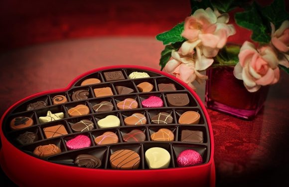 A heart-shaped box of assorted chocolates next to a pink vase with pink and white flowers on a dark wooden surface.