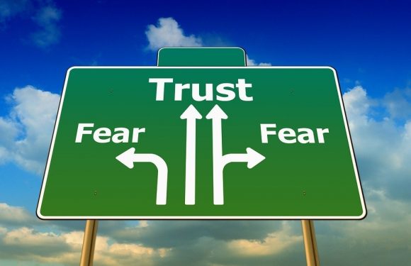 A green road sign with white arrows and text, showing three options: left for fear, straight for trust, and right for fear, against a cloudy sky background.