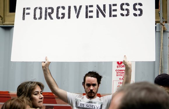 A man holding a large sign with the word "FORGIVENESS" written on it, while wearing a t-shirt that says "I'm sorry." Several people are in the foreground.