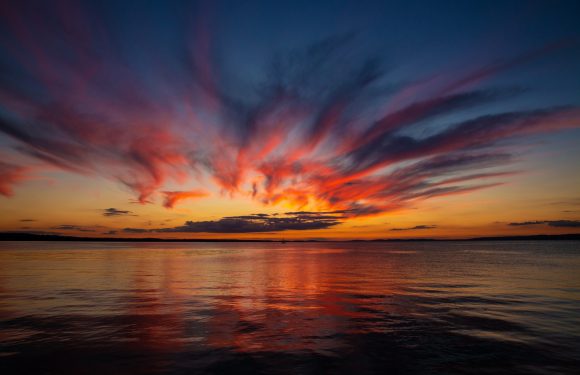 A vibrant sunset over a calm body of water, with colorful clouds in shades of orange, pink, and purple reflecting on the surface.