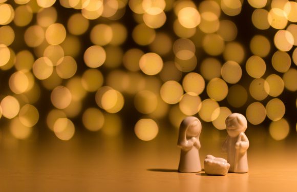 Small ceramic nativity figures of Mary and Joseph with baby Jesus on a surface, with warm, blurred golden lights in the background.