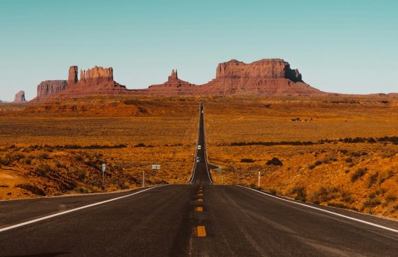 A long straight road extends through a desert landscape with red rock formations in the distance under a clear blue sky.