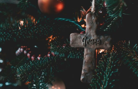 A close-up of a Christmas tree adorned with ornaments, including a wooden cross with "Grace" inscribed on it, surrounded by green pine branches and red berries.