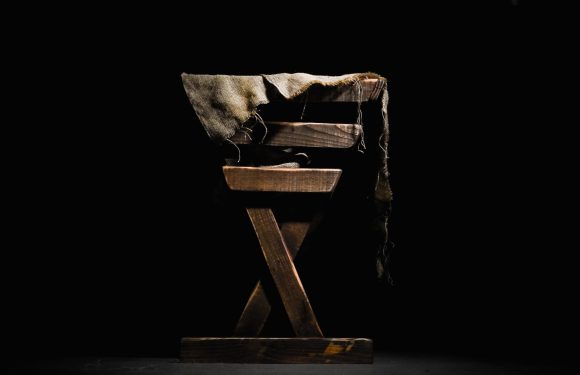 A worn wooden chair with a frayed, distressed piece of fabric draped over the top, set against a black background with light highlighting the chair's texture.