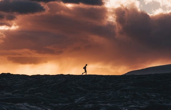 A person hiking on a rocky terrain at sunset, with a dramatic sky filled with dark clouds and glowing light near the horizon.