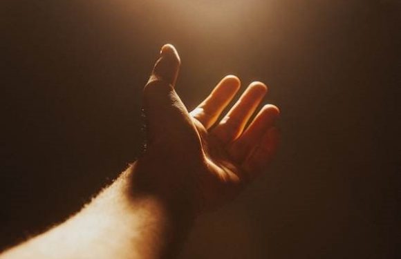 A hand reaching upward toward a bright light source with a plain, dark background, creating a silhouette effect.