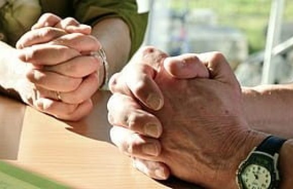 Two pairs of hands clasped in prayer over a table, with a person wearing a watch and a striped shirt on the right and others on the left.