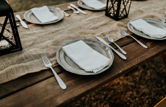 A rustic outdoor table set with white plates, silverware, napkins, black lanterns, and a burlap table runner for a dinner event.