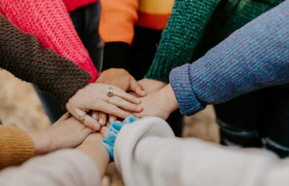 Multiple hands stacked together in the center, with some individuals wearing colorful sweaters and one with a blue scrunchie on their wrist.