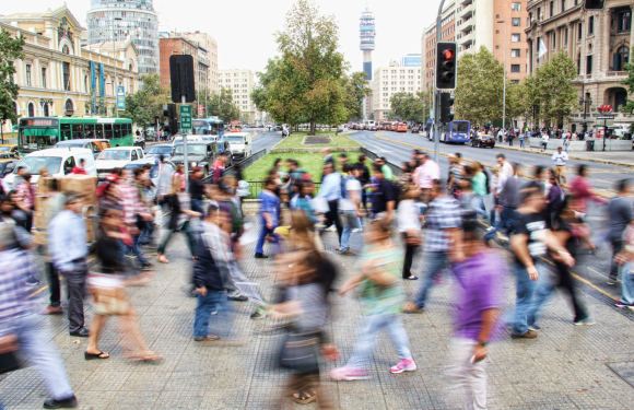 A large crowd of blurred pedestrians crosses a busy city intersection with traffic lights, surrounded by tall buildings and trees, and a city tower visible in the background.