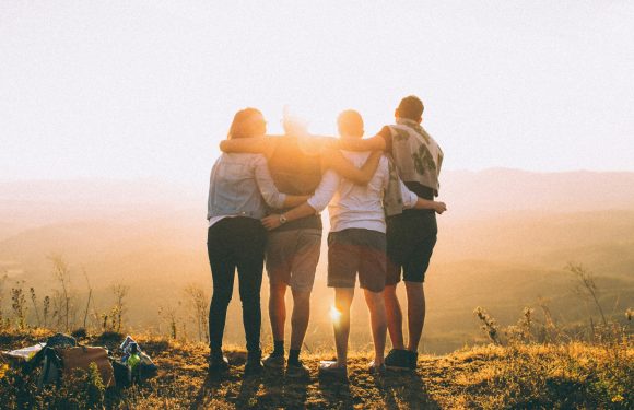 Group of five friends standing close together with arms around each other, enjoying a sunset on a grassy hilltop. They are casually dressed with picnic items nearby.