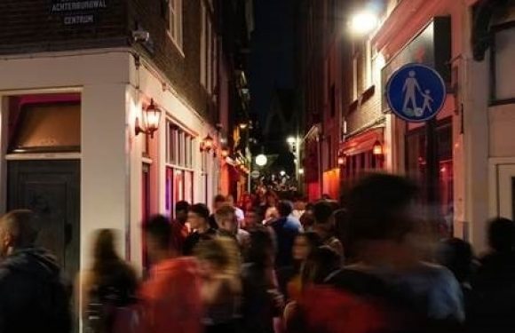 Crowded narrow street at night with people walking, warm lighting, and a sign indicating a pedestrian walkway. A building with a red exterior is on the right.