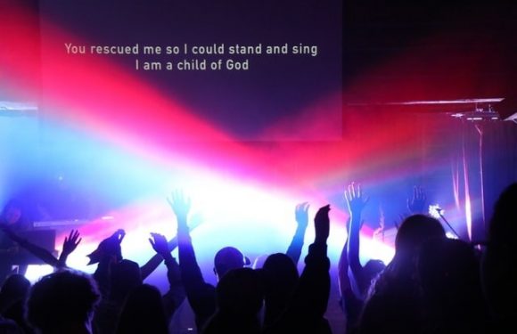 People raising hands during a concert or worship event with a bright light and a screen displaying the lyrics "You rescued me so I could stand and sing I am a child of God."