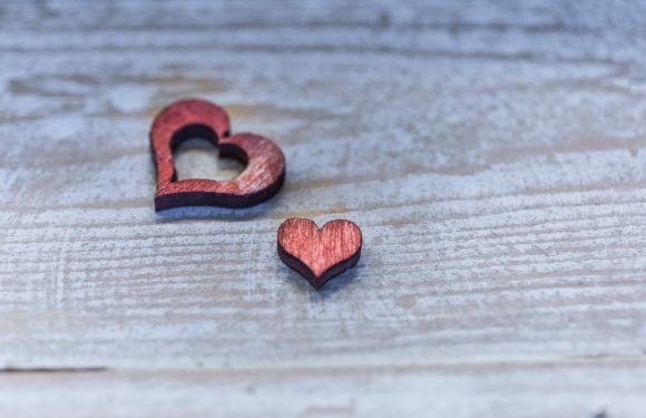Two small wooden hearts, one larger with a hollow center, on a light wooden surface with visible grain. The hearts are reddish with dark edges.