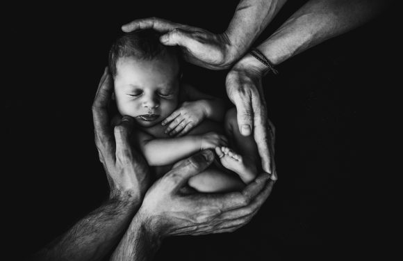 Black-and-white image of a tiny newborn baby cradled and surrounded by multiple hands, conveying tenderness and protection. The baby appears peaceful and asleep.