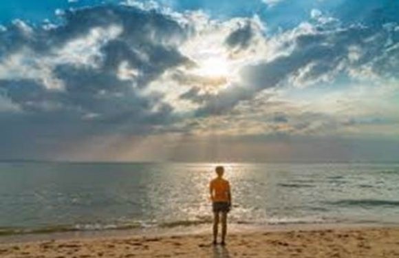 A person in an orange shirt and shorts stands on the sandy beach, looking at the ocean under a partly cloudy sky with the sun shining through clouds.